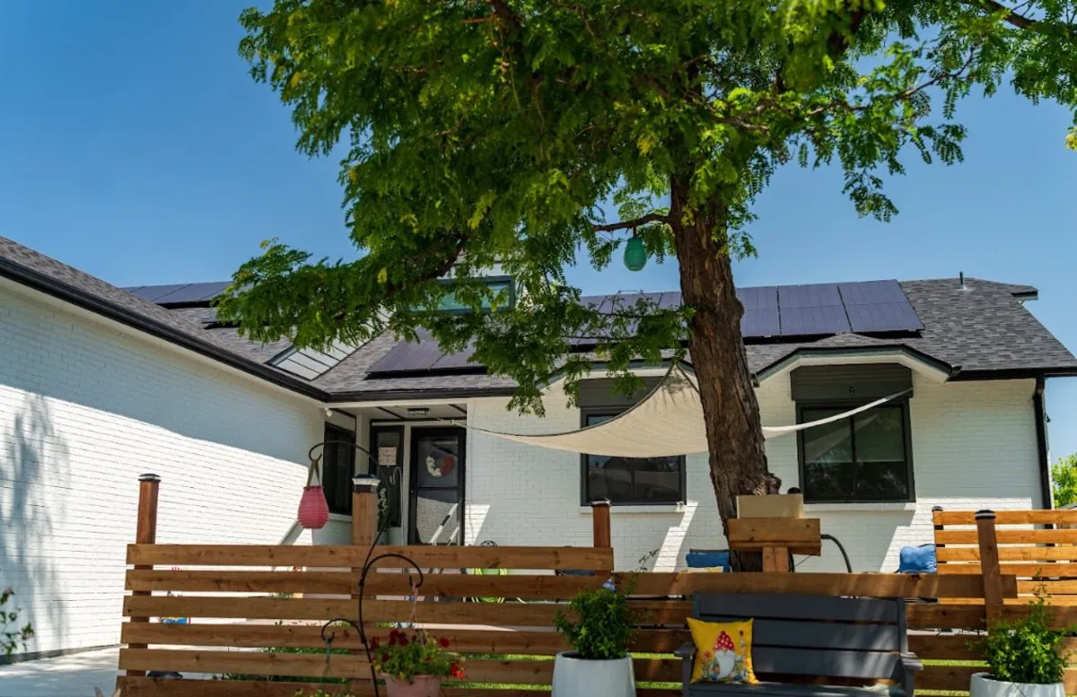 White brick Colorado home with a black rooftop solar array, shaded by a mature tree
